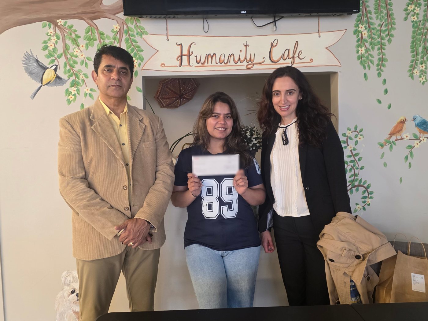 Amrin holding the Vancity grant cheque inside the Humanity Café, with two guests